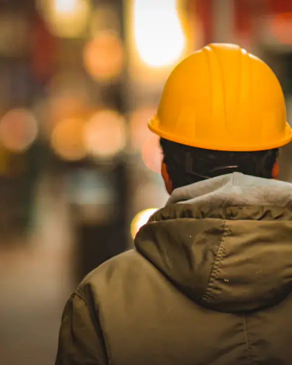 Beispielbild: Arbeiter mit gelbem Schutzhelm von hinten, im Hintergrund unscharfe Stadtlichter