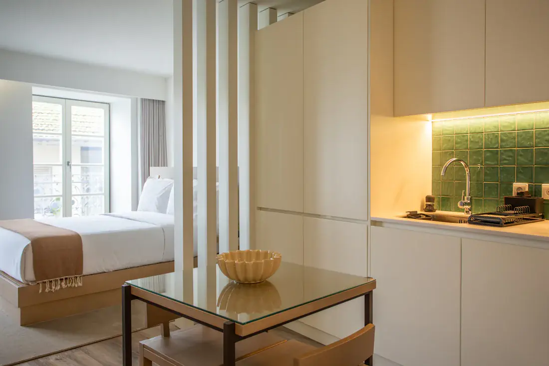 Kompaktes Studio mit Küchenzeile und Glastisch, dahinter das Bett am Fenster