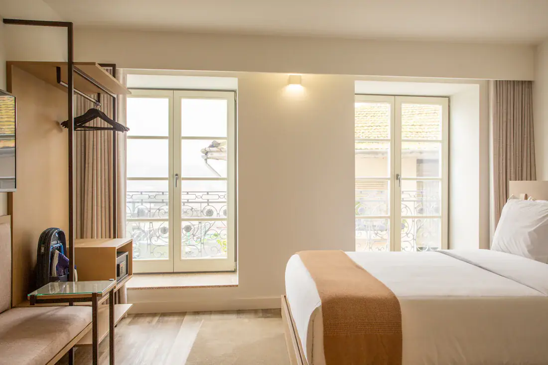 Beispielbild: helles Studio mit Doppelbett am bodentiefen Fenster, Holzboden und weichem Tageslicht